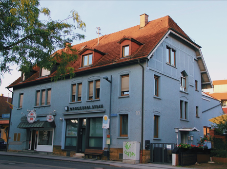 Ein längliches hellblaues Haus mit rot-braunem Dach, an der Fassade der Schriftzug "Druckerei Stein".