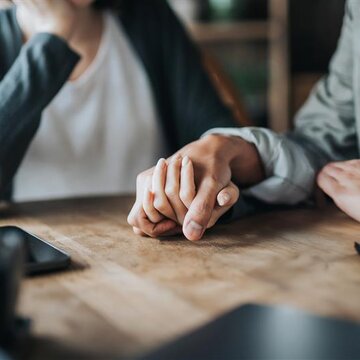 Zwei Personen sitzen am Tisch und halten die Hand der anderen Person.