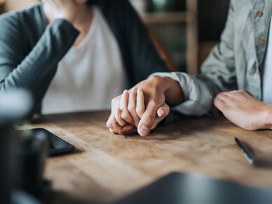 Zwei Personen sitzen am Tisch und halten die Hand der anderen Person.