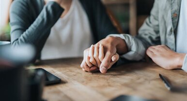 Zwei Personen sitzen am Tisch und halten die Hand der anderen Person.